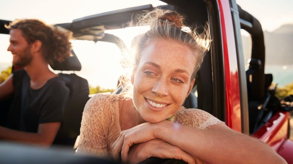Millennial white couple on a road trip driving in open top car, women leaning on car door, close up