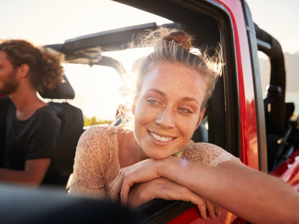 Millennial white couple on a road trip driving in open top car, women leaning on car door, close up